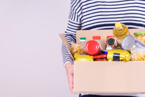 Volunteer hands holding food donations box with grocery products on white desk.