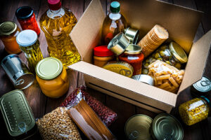 High angle view of a cardboard box filled with multicolored non-perishable canned goods, conserves, sauces and oils shot on wooden table. The composition includes cooking oil bottle, pasta, crackers, preserves and tins. High resolution 42Mp studio digital capture taken with SONY A7rII and Zeiss Batis 40mm F2.0 CF lens