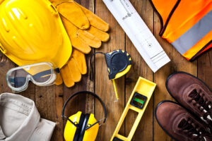 Personal protective workwear and blueprint with some measuring instruments shot directly from above on rustic wood background. The protective workwear includes hard hat, gloves, earmuff, goggles, steel toe shoes, and safety vest. The composition also includes a tape measure and bubble level and a construction blueprint, all items used by construction worker or engineer. Predominant colors: yellow and brown.